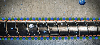 Overhead view of industrial metal pipe with blue and green spheres arranged along its length on concrete surface