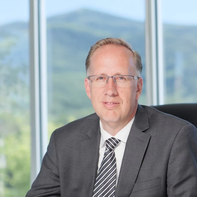 Professional headshot of male executive in gray suit and striped tie against mountain landscape window backdrop Headshot
