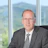 Professional headshot of male executive in gray suit and striped tie against mountain landscape window backdrop Headshot