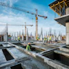 Construction workers in safety vests on concrete foundation with cranes and steel framework at active building site