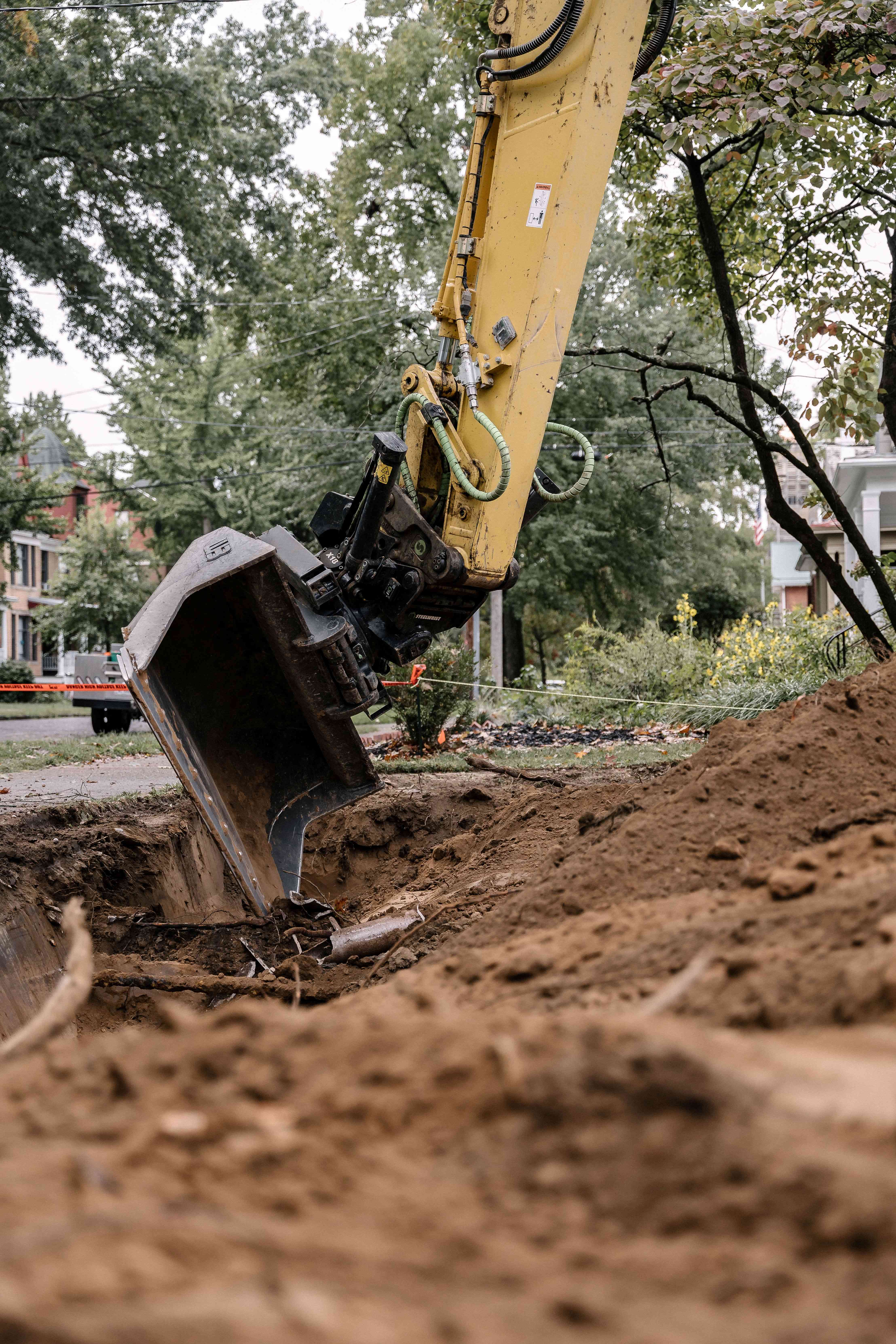 Yellow excavator digging soil at urban construction site surrounded by trees and buildings