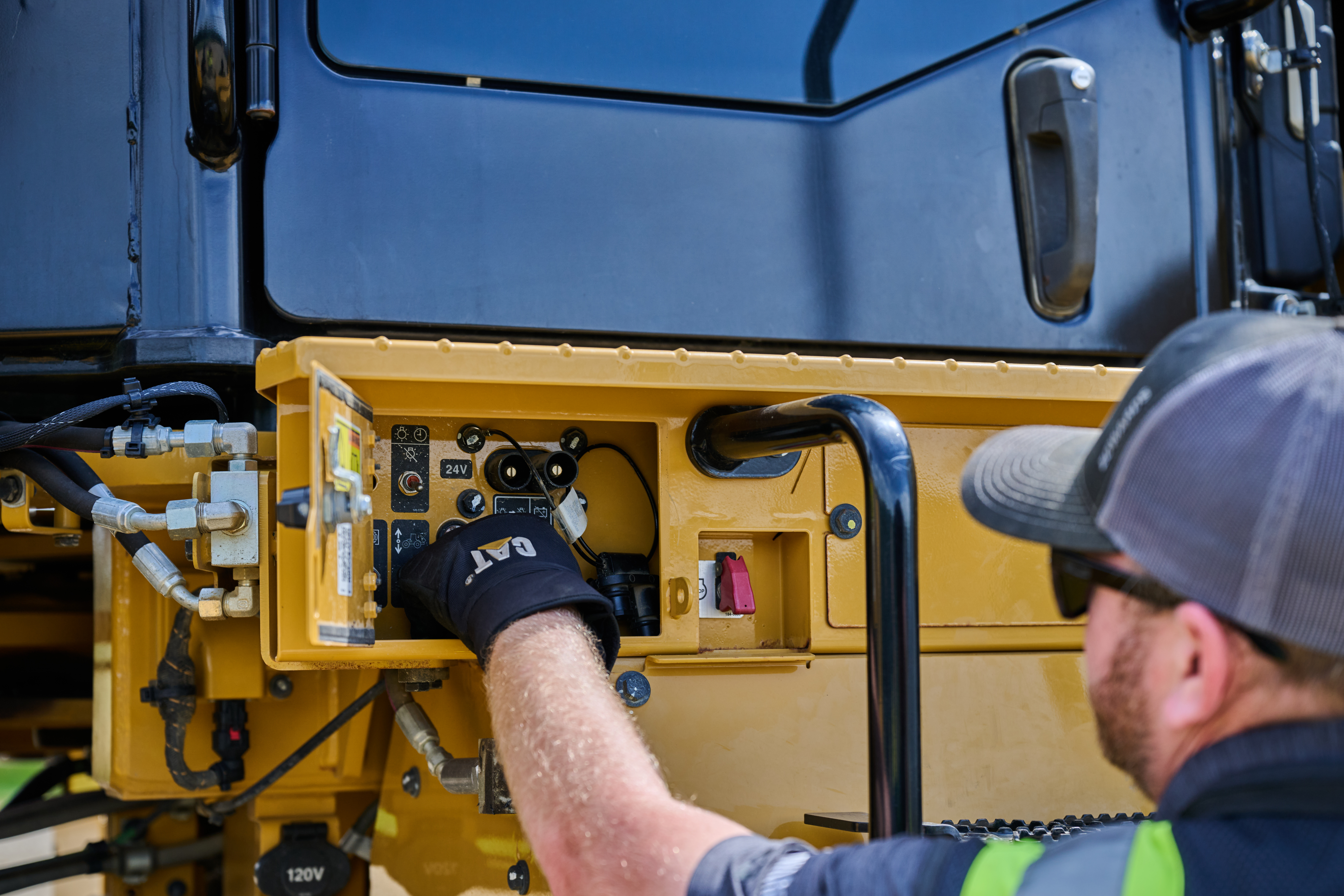 Technician in safety gear inspecting yellow heavy equipment control panel with blue cab visible above