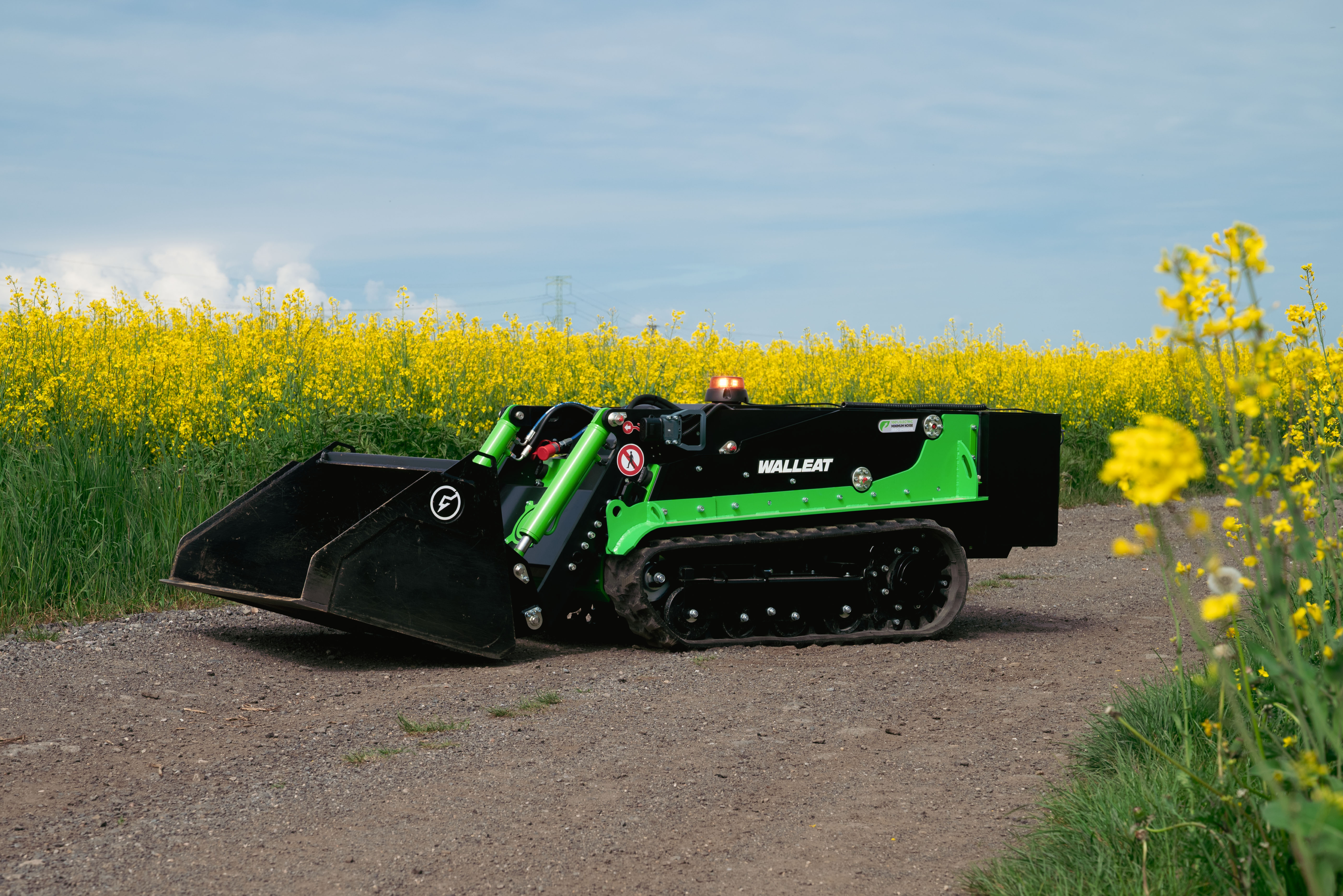 Green and black tracked mini loader positioned on a rural road with yellow rapeseed field in background