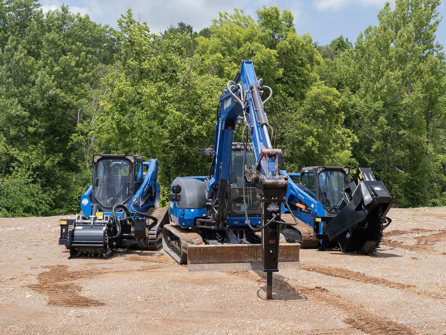Blue construction equipment including excavator and skid steer loaders positioned at a dirt job site with trees in background