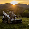 Bobcat skid steer loader positioned in grassy field at sunset with forested mountains in background