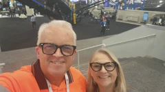 Two people smiling at an industrial trade show with machinery and equipment visible in the background
