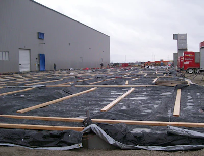 Industrial roofing construction site with black membrane sheets, wooden supports, warehouse building, and red truck in background