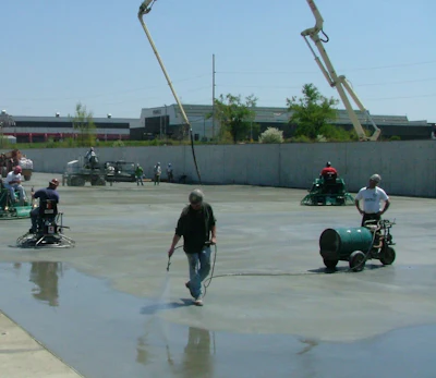 People gathered on wet concrete surface with vintage tractors and large metal sculptural structures overhead at outdoor event