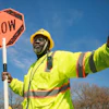 Construction worker in safety gear holding an orange SLOW traffic sign against blue sky