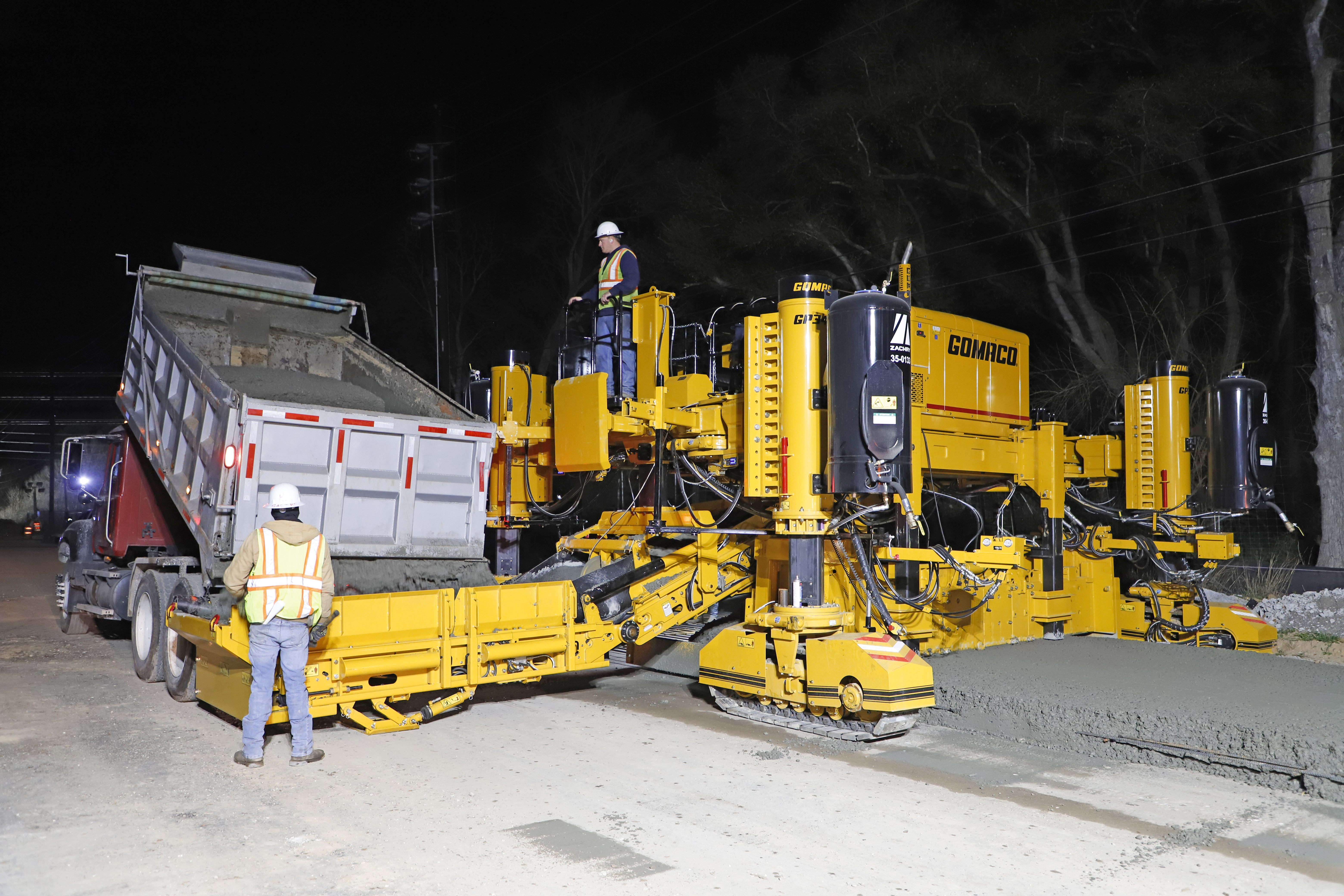 Gomaco concrete paving machine operating at night with workers and dump truck during road construction.