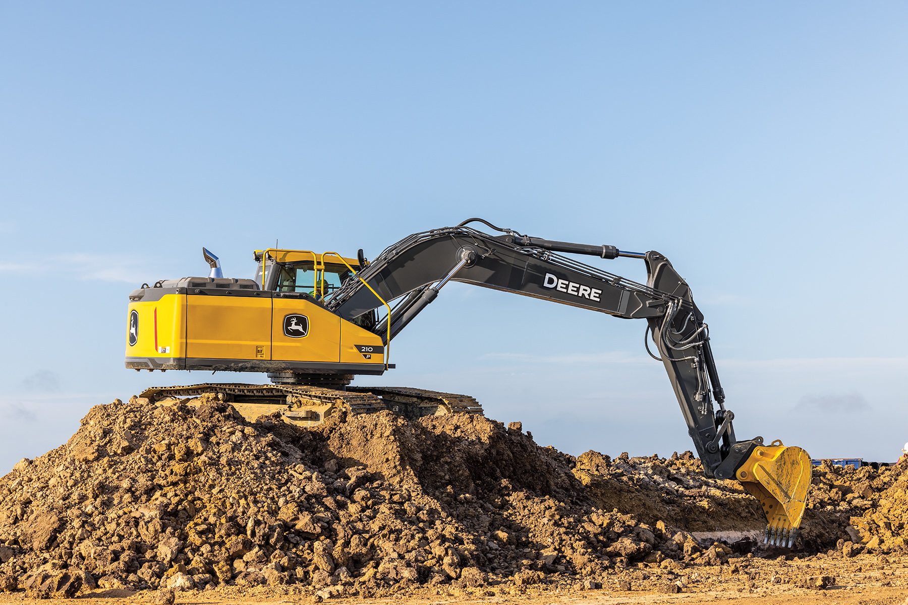 Yellow John Deere excavator on a dirt pile against a clear blue sky