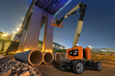 Orange JLG telescopic boom lift operating at dusk near large industrial pipes and concrete structure