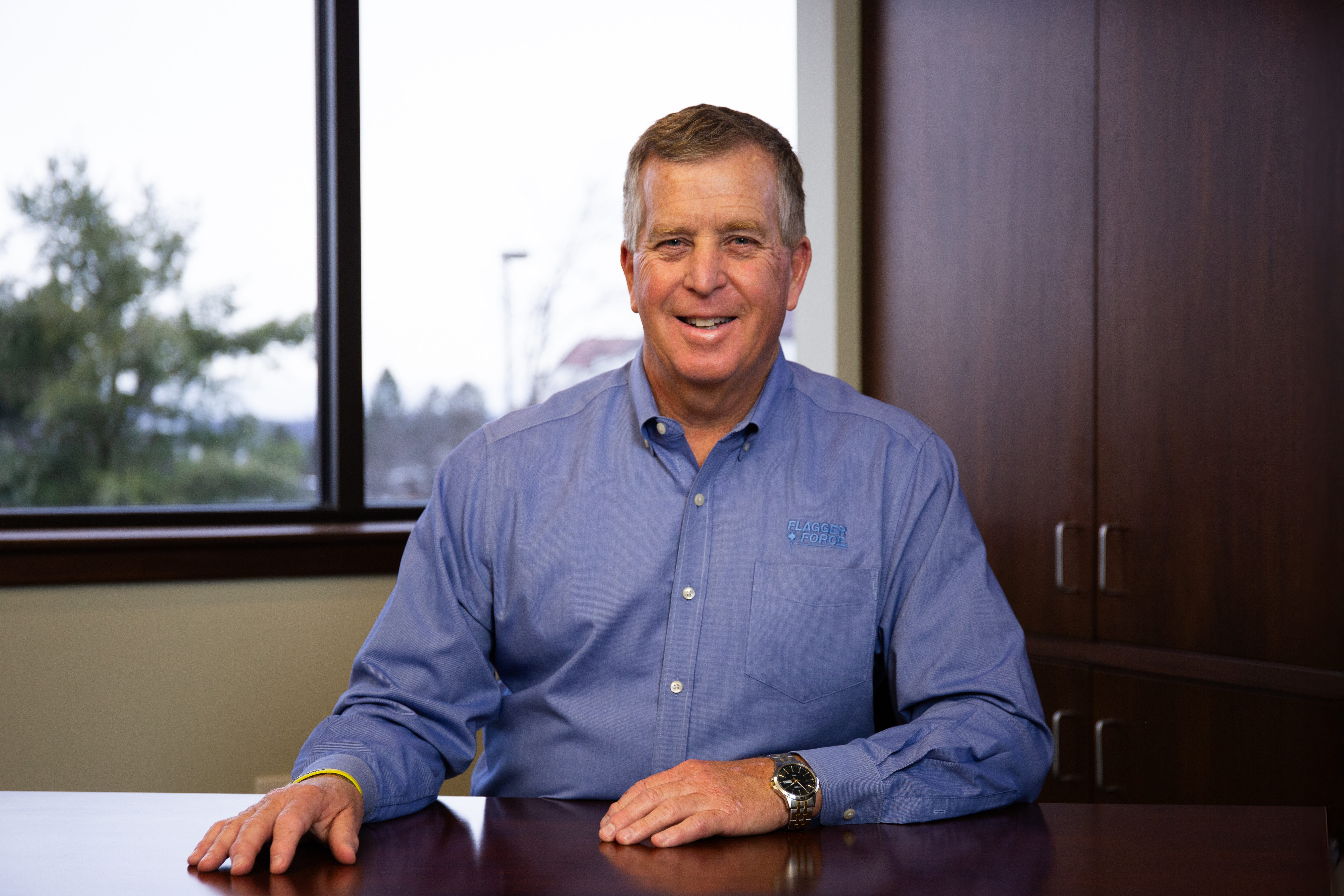 Professional man in blue button-up shirt smiling at desk with window view behind him Headshot