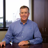 Professional man in blue button-up shirt smiling at desk with window view behind him