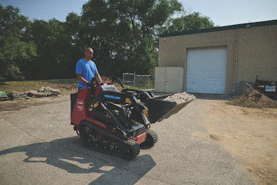 Man operating mini skid steer loader at industrial facility with metal building
