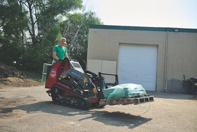 Worker operating red tracked mini loader with green wrapped material at industrial facility