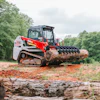 Red and white Bobcat compact track loader with grapple attachment holding large log in wooded area