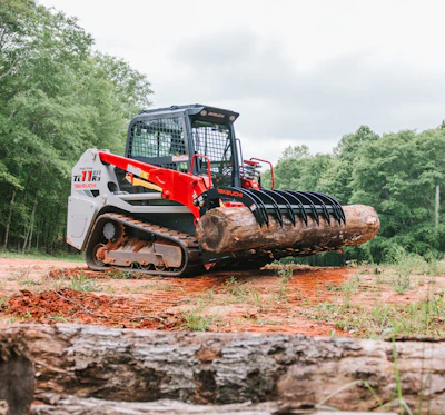 Red and white Bobcat compact track loader with grapple attachment holding large log in wooded area