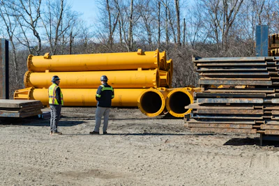Two workers in safety vests inspect large yellow industrial pipes at a construction yard with bare trees and stacked materials in background