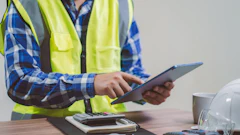 Construction worker in safety vest using tablet at desk with coffee cup and supplies