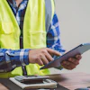 Construction worker in safety vest using tablet at desk with coffee cup and supplies