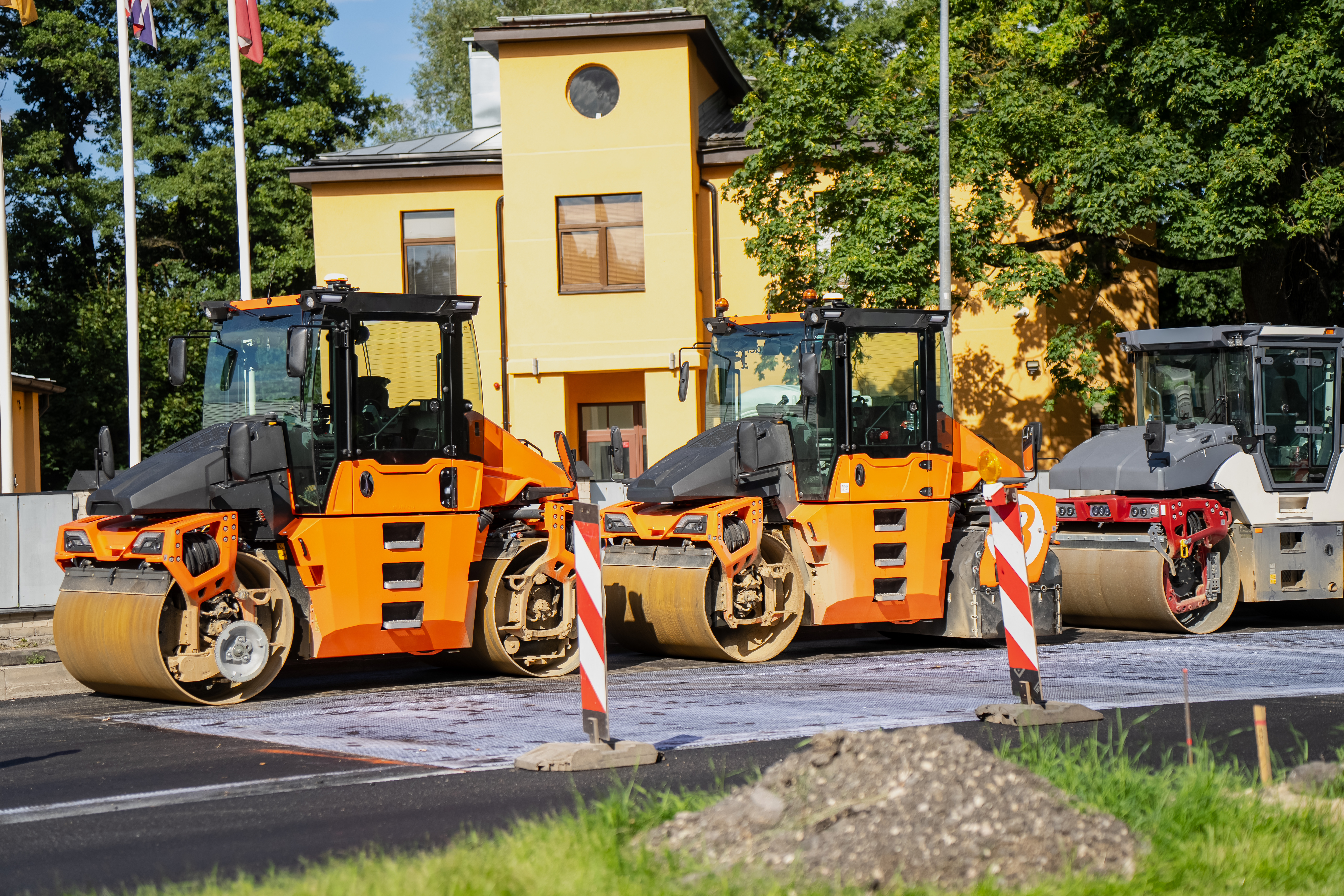 Three orange and black road compactor rollers lined up at a construction site with yellow building in background