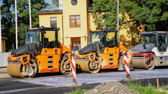 Three orange and black road compactor rollers lined up at a construction site with yellow building in background