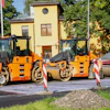 Three orange and black road compactor rollers lined up at a construction site with yellow building in background