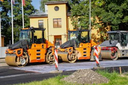 Three orange and black road compactor rollers lined up at a construction site with yellow building in background