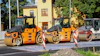 Three orange and black road compactor rollers lined up at a construction site with yellow building in background