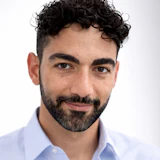Professional headshot of a man with dark curly hair and beard wearing a light blue button-up shirt against a white background