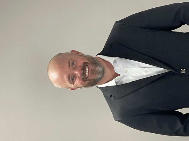 Professional headshot of a smiling man in dark blazer and white shirt against neutral background