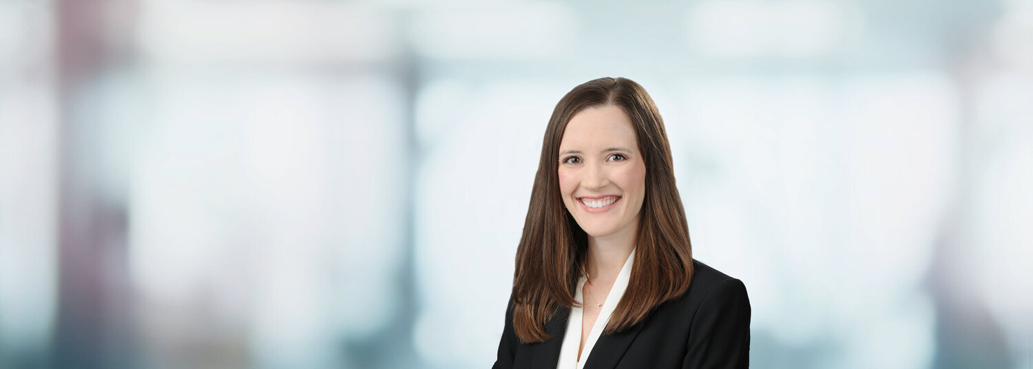 Professional headshot of woman in black blazer and white blouse smiling against blurred office background