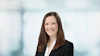 Professional headshot of woman in black blazer and white blouse smiling against blurred office background