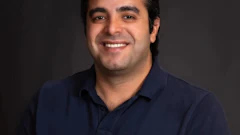 Professional headshot of a man in navy polo shirt smiling against dark background