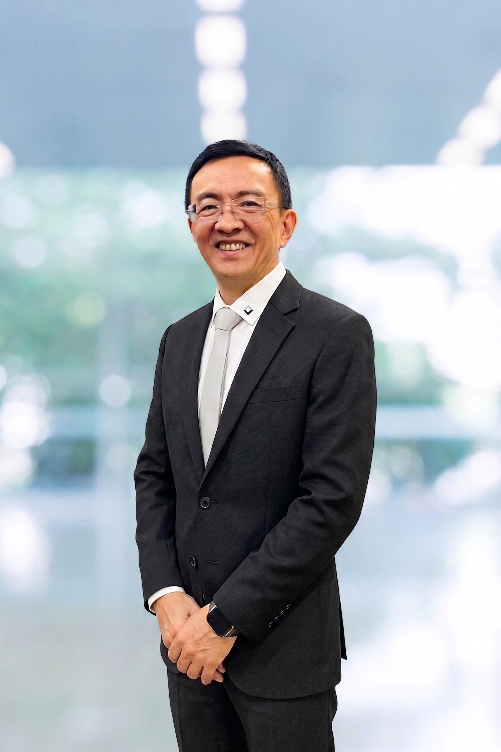 Professional business headshot of an Asian man in black blazer and glasses smiling against blurred cityscape background
