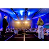 Two business speakers present on stage to an audience in a modern conference hall with blue ambient lighting and warm spotlights.