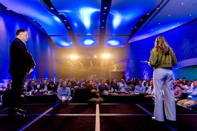 Two business speakers present on stage to an audience in a modern conference hall with blue ambient lighting and warm spotlights.
